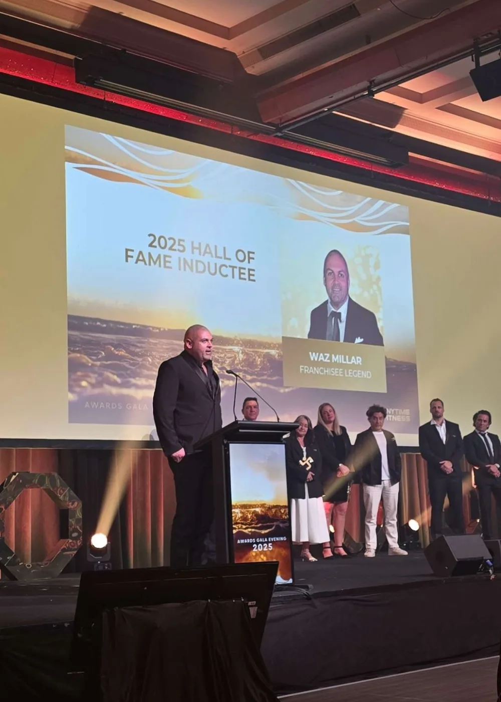 Waz Millar speaking on stage at the 2025 Anytime Fitness awards gala, with the screen behind him naming him a Hall of Fame inductee.